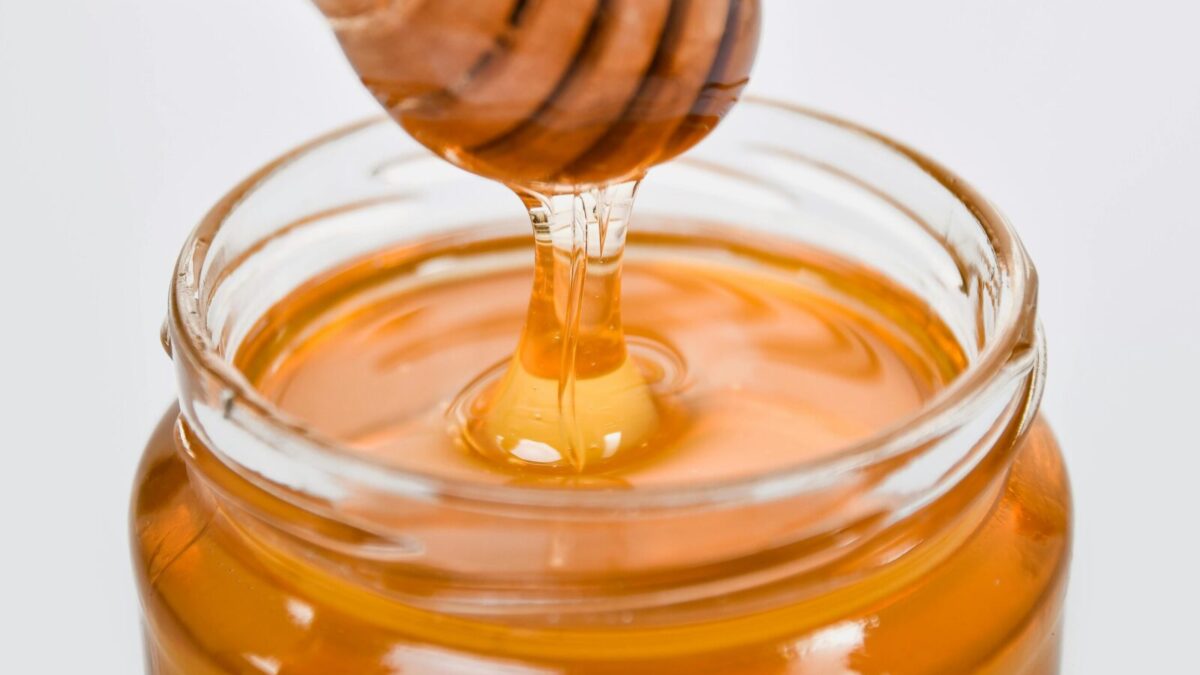 Close-up of honey flowing from a wooden dipper into a glass jar on a white background.