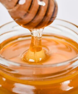 Close-up of honey flowing from a wooden dipper into a glass jar on a white background.