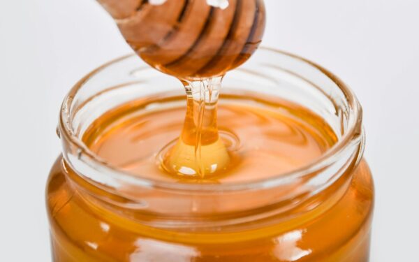 Close-up of honey flowing from a wooden dipper into a glass jar on a white background.