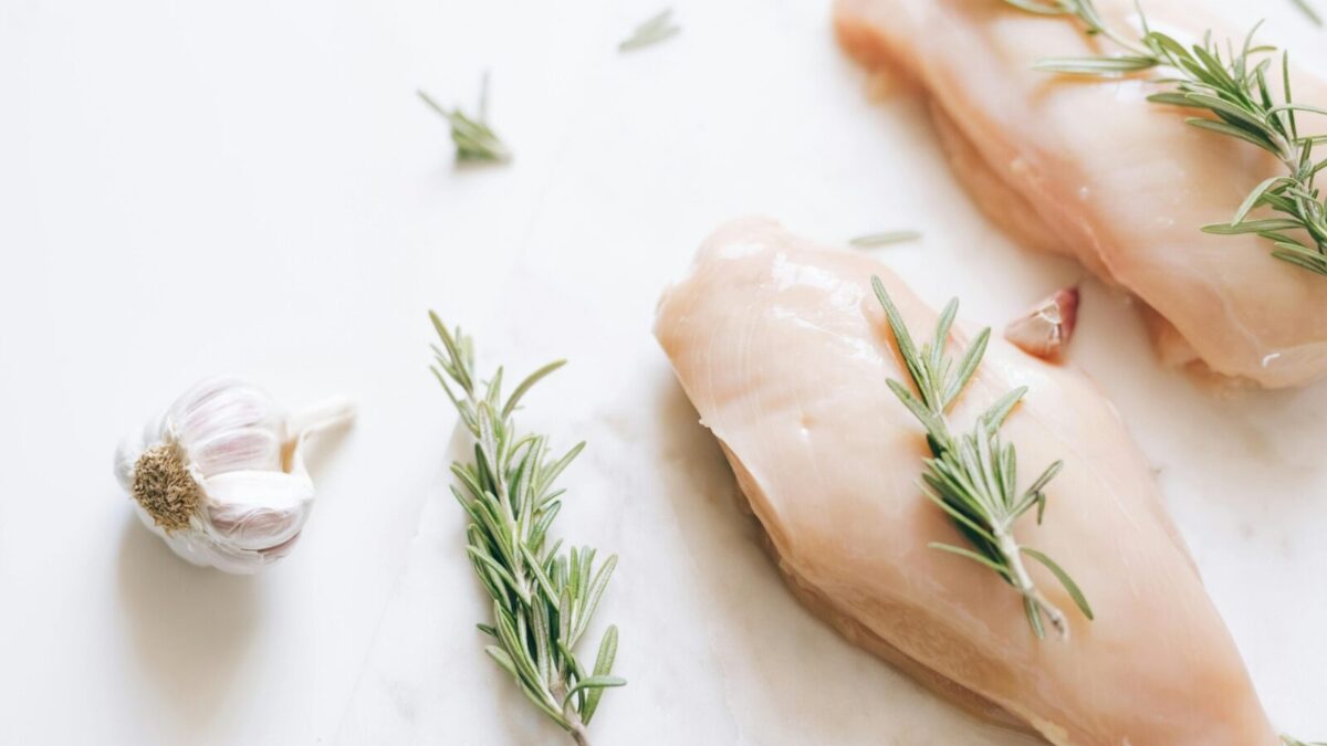 Raw chicken breasts garnished with rosemary and garlic on a white surface.