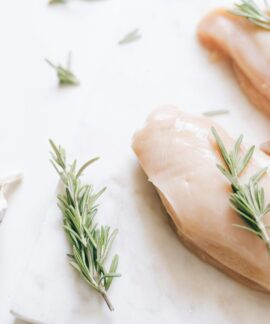Raw chicken breasts garnished with rosemary and garlic on a white surface.