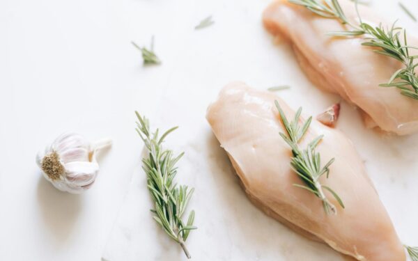 Raw chicken breasts garnished with rosemary and garlic on a white surface.