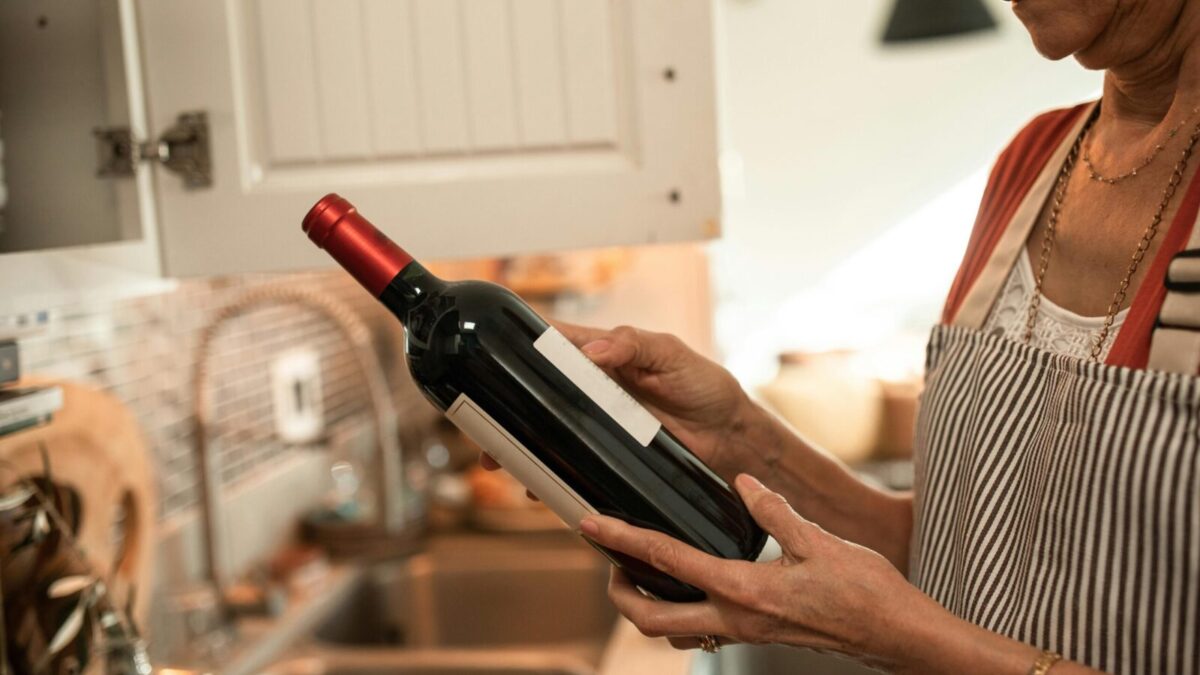 A senior woman examines a wine bottle in her cozy kitchen, wearing a striped apron.
