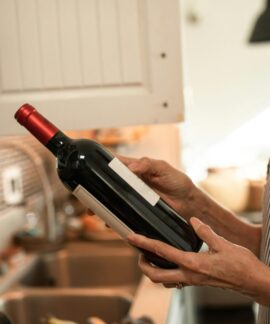 A senior woman examines a wine bottle in her cozy kitchen, wearing a striped apron.