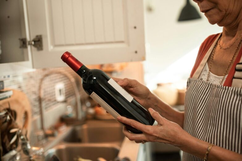 A senior woman examines a wine bottle in her cozy kitchen, wearing a striped apron.