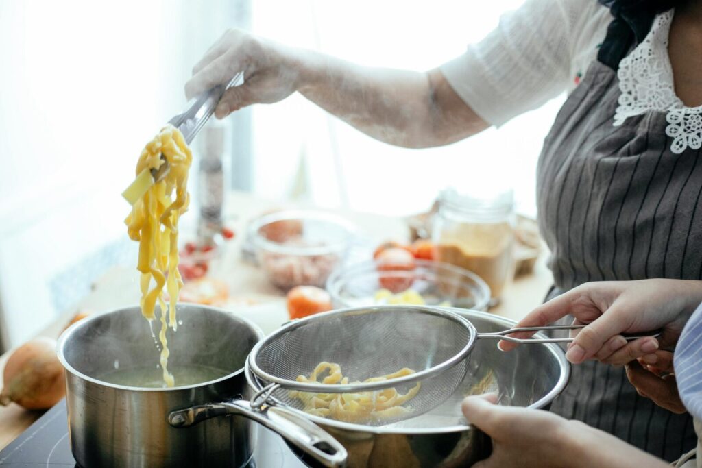 Crop anonymous females in aprons cooking delicious pasta using tongs and strainer in kitchen