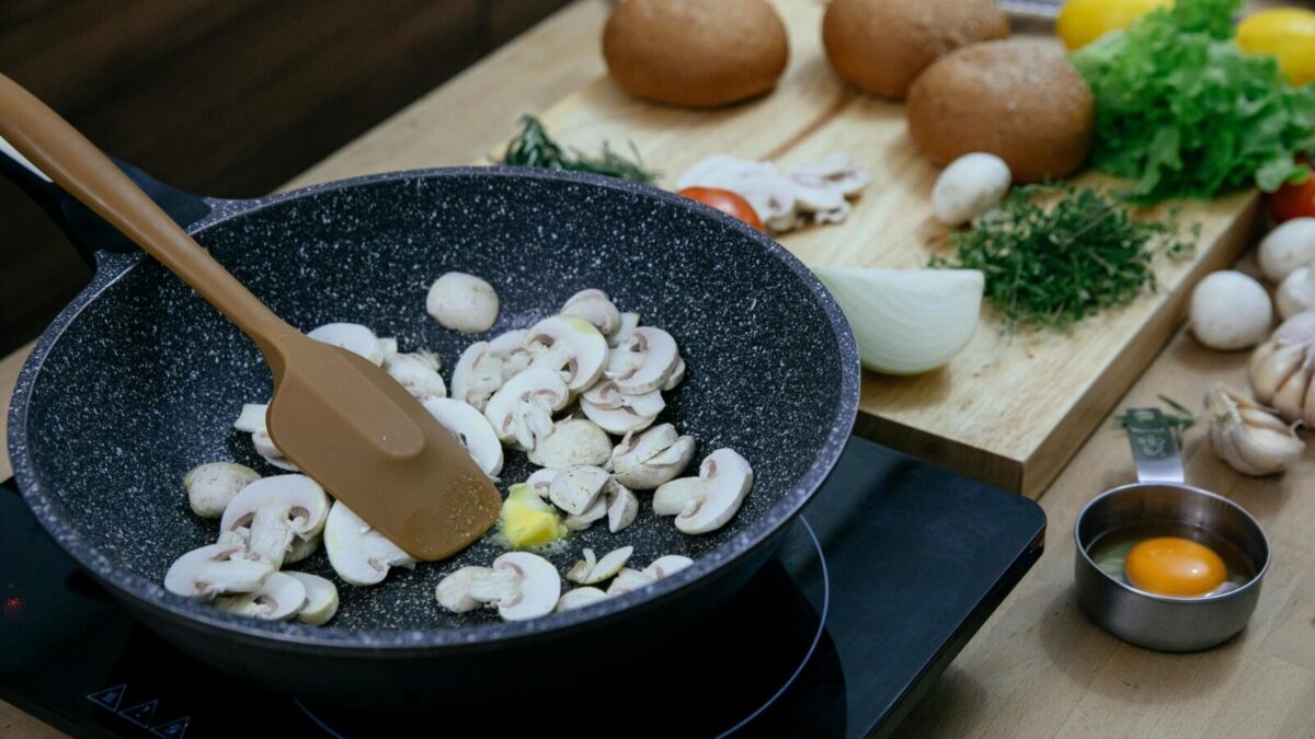 From above of silicone spatula in pan with frying chopped mushrooms placed on electric stove near wooden cutting board with assorted herbs and vegetables in kitchen