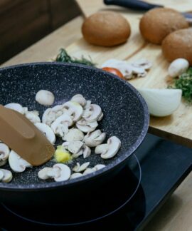 From above of silicone spatula in pan with frying chopped mushrooms placed on electric stove near wooden cutting board with assorted herbs and vegetables in kitchen