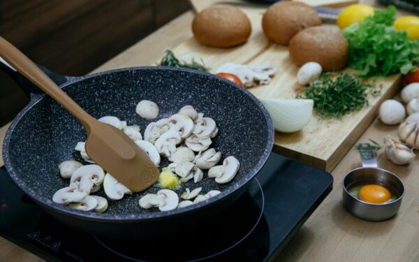 From above of silicone spatula in pan with frying chopped mushrooms placed on electric stove near wooden cutting board with assorted herbs and vegetables in kitchen