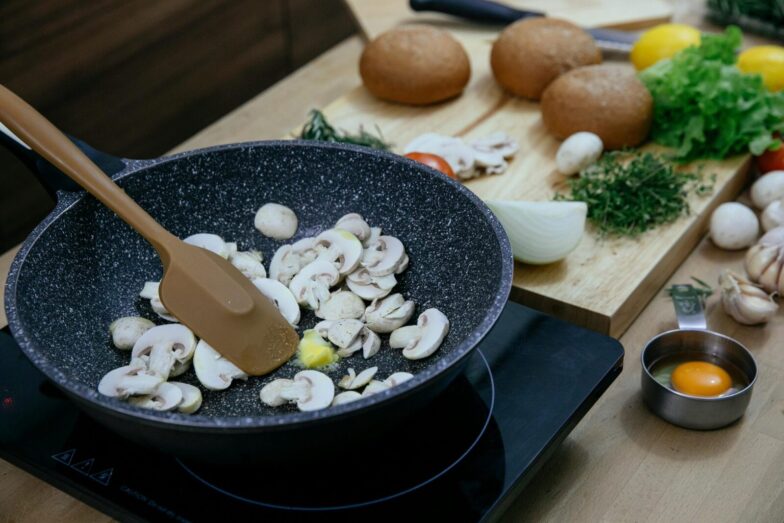 From above of silicone spatula in pan with frying chopped mushrooms placed on electric stove near wooden cutting board with assorted herbs and vegetables in kitchen