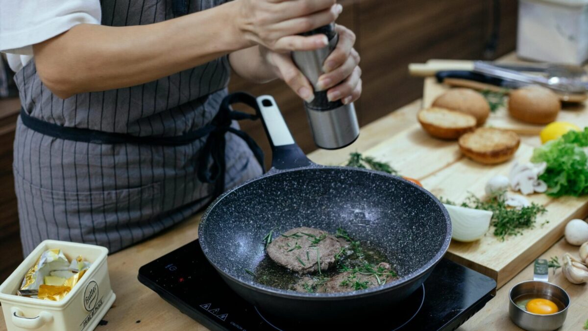 Unrecognizable female cook wearing apron adding seasoning on cutlets while preparing delicious burger at table with stove in kitchen
