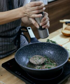 Unrecognizable female cook wearing apron adding seasoning on cutlets while preparing delicious burger at table with stove in kitchen