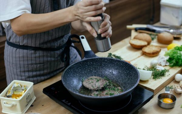 Unrecognizable female cook wearing apron adding seasoning on cutlets while preparing delicious burger at table with stove in kitchen