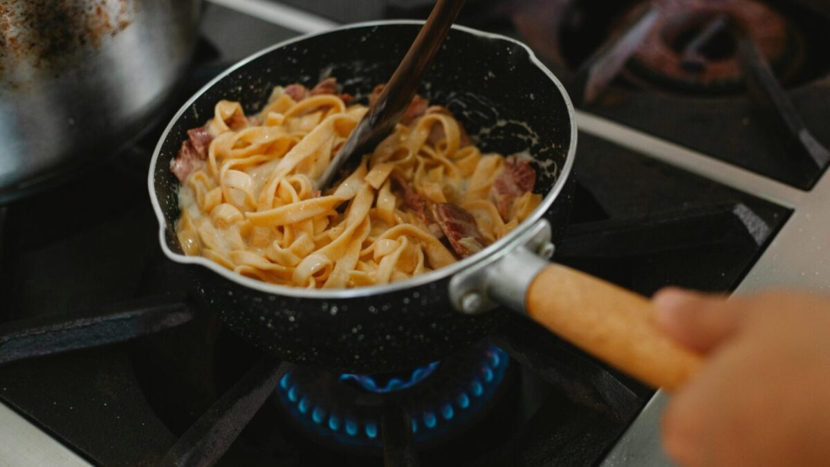A close-up shot of pasta with bacon being cooked in a frying pan on a stove.