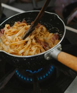 A close-up shot of pasta with bacon being cooked in a frying pan on a stove.