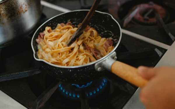 A close-up shot of pasta with bacon being cooked in a frying pan on a stove.