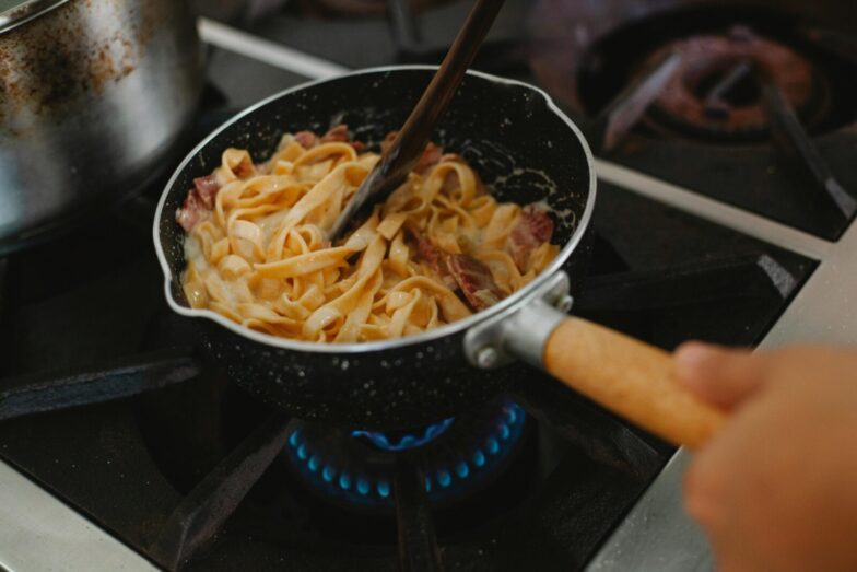 A close-up shot of pasta with bacon being cooked in a frying pan on a stove.