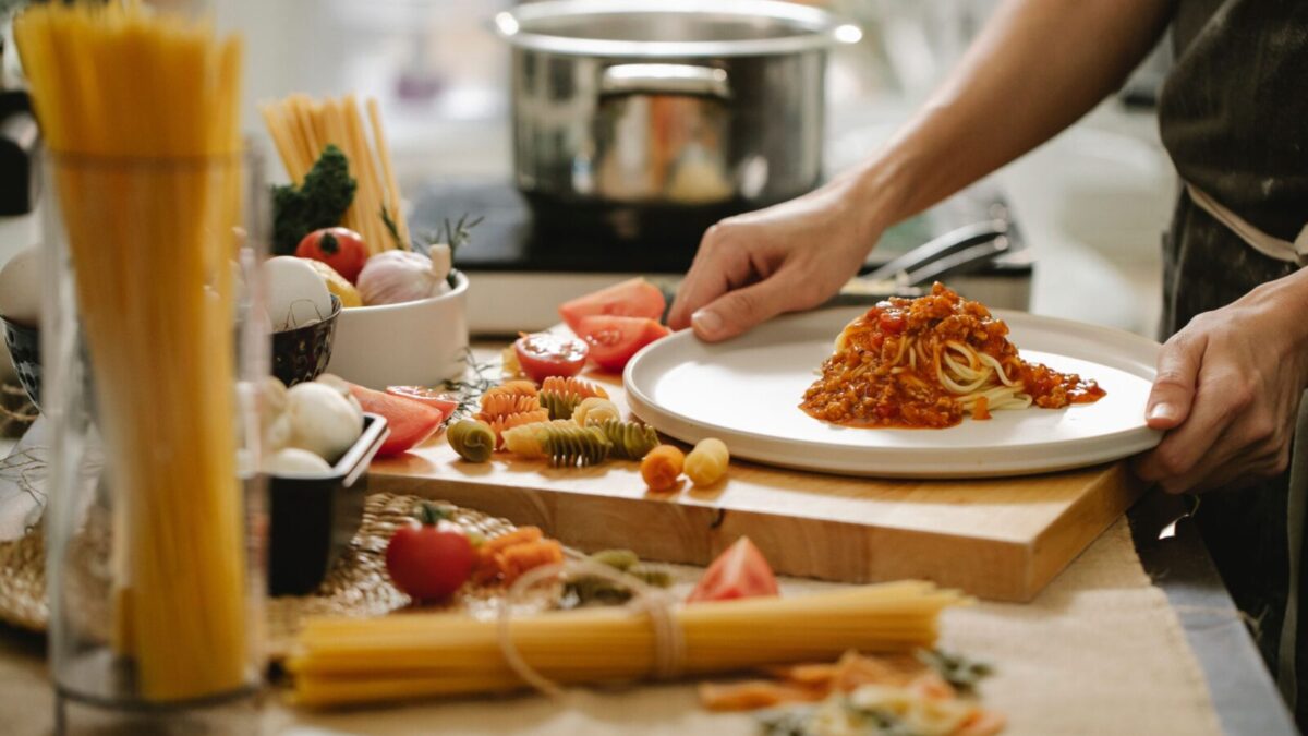 Crop anonymous cook standing at table with various ingredients and cooking pasta with meat and tomatoes