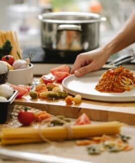 Crop anonymous cook standing at table with various ingredients and cooking pasta with meat and tomatoes