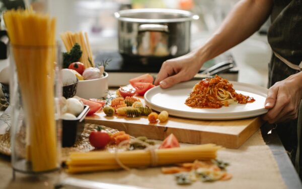 Crop anonymous cook standing at table with various ingredients and cooking pasta with meat and tomatoes
