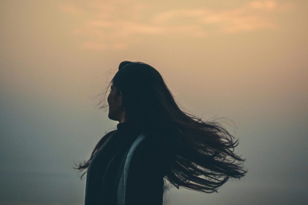 Side view of unrecognizable female shaking long hair while standing on shore near foggy sea on evening time in nature