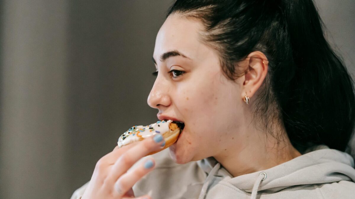 Crop young brunette in comfy hoodie eating fresh appetizing doughnut covered with vanilla icing and sprinkles in light room