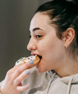 Crop young brunette in comfy hoodie eating fresh appetizing doughnut covered with vanilla icing and sprinkles in light room