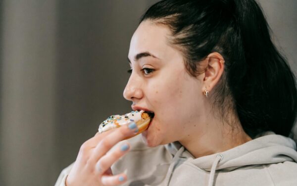 Crop young brunette in comfy hoodie eating fresh appetizing doughnut covered with vanilla icing and sprinkles in light room