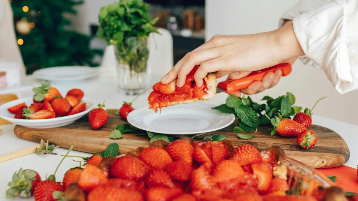 A hand slicing fresh homemade strawberry pie in a kitchen setting, surrounded by vibrant strawberries and mint.