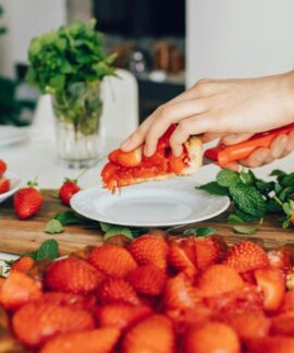 A hand slicing fresh homemade strawberry pie in a kitchen setting, surrounded by vibrant strawberries and mint.