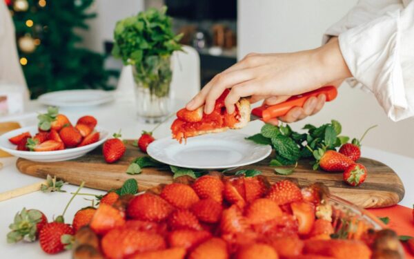 A hand slicing fresh homemade strawberry pie in a kitchen setting, surrounded by vibrant strawberries and mint.