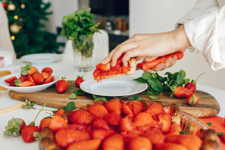 A hand slicing fresh homemade strawberry pie in a kitchen setting, surrounded by vibrant strawberries and mint.