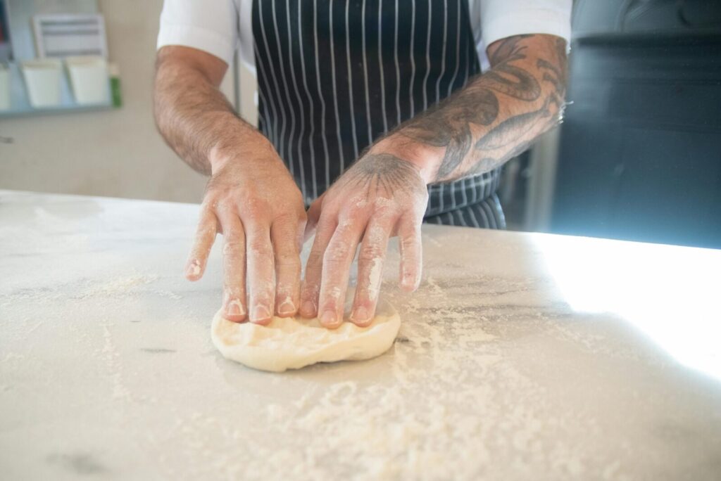 A chef with tattoos kneads dough on a floured surface in a kitchen setting.