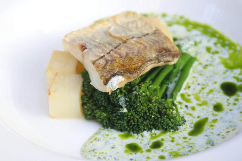 Piece of tasty grilled fish with fresh green broccoli served on white round plate with sauce and vegetable on table