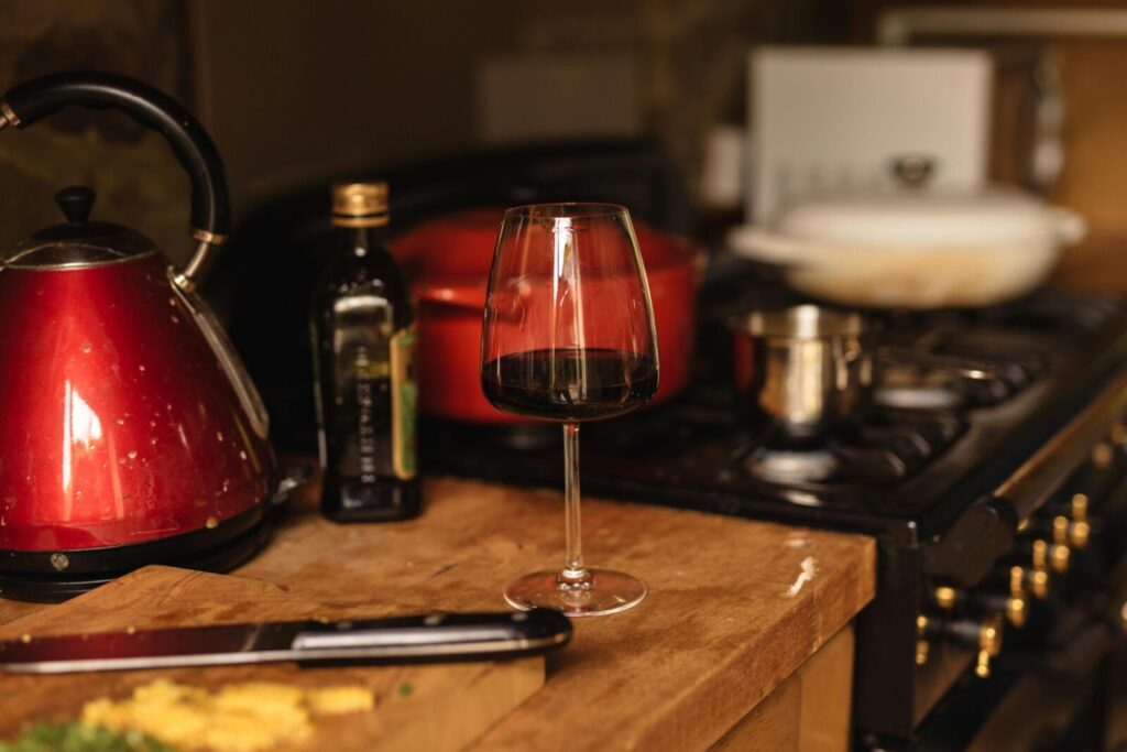 Elegant crystal glass of red wine placed on wooden table in kitchen near stove and various utensils at home