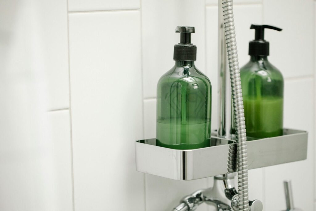 Green dispensers with body lotion on shelf on shower system in bathroom against tiled wall