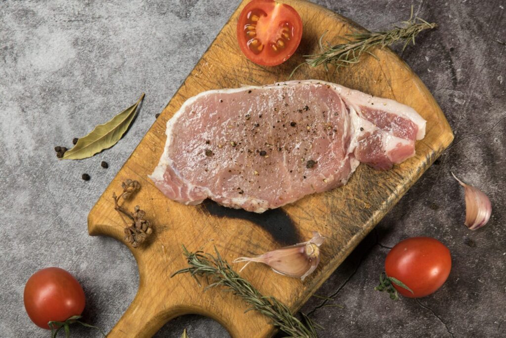 Top view of raw pork steak on a wooden board with fresh herbs and tomatoes.