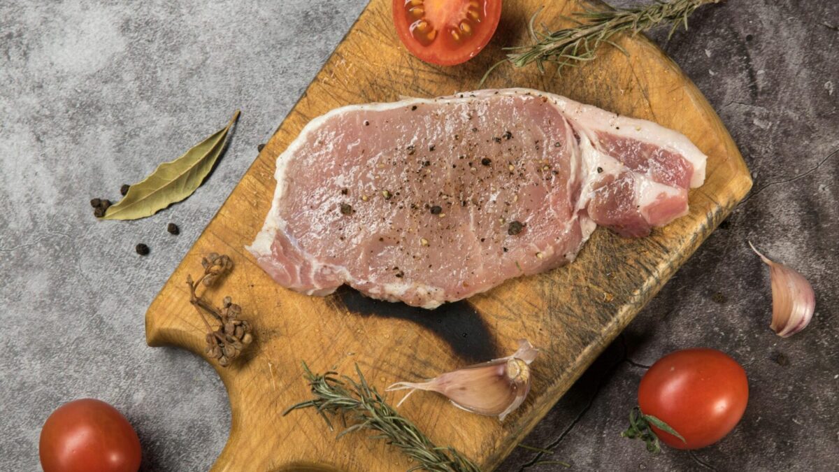 Top view of raw pork steak on a wooden board with fresh herbs and tomatoes.