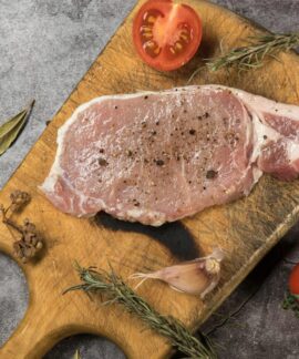 Top view of raw pork steak on a wooden board with fresh herbs and tomatoes.