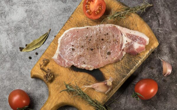 Top view of raw pork steak on a wooden board with fresh herbs and tomatoes.