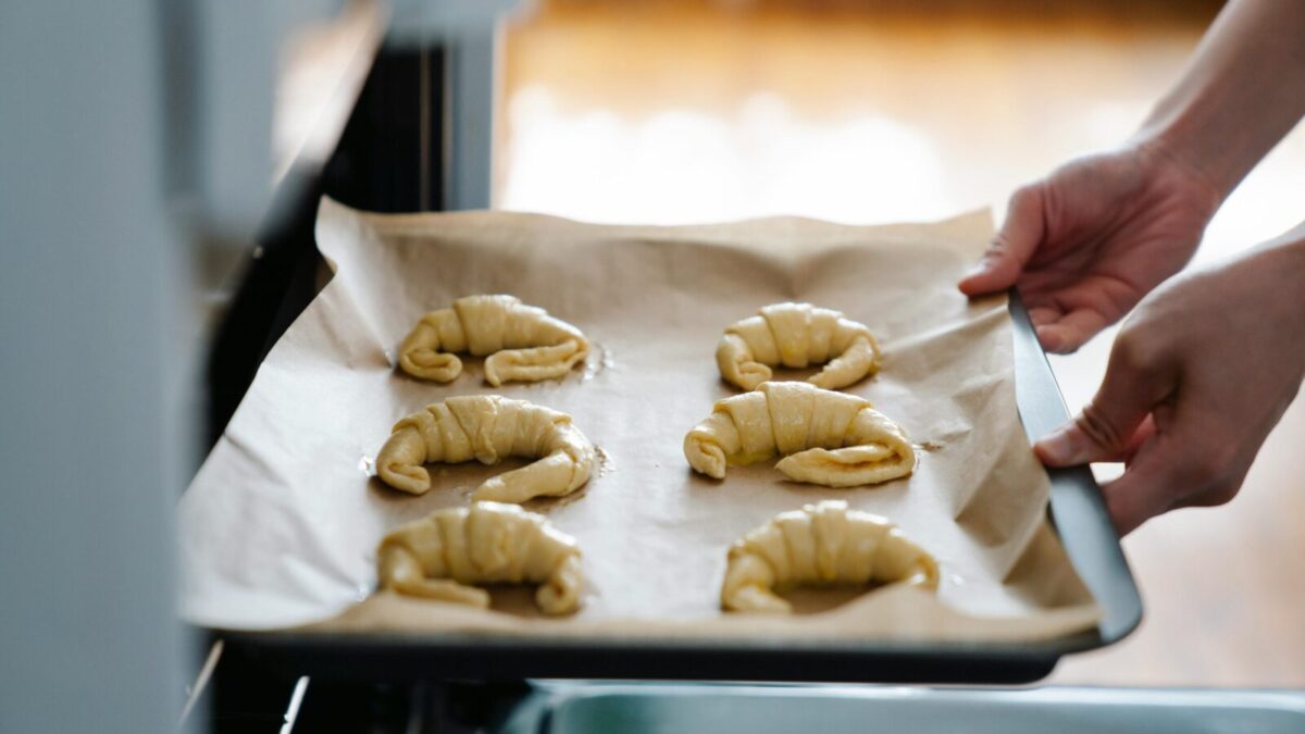 Hands placing homemade croissants on a tray into an oven, ready to bake.