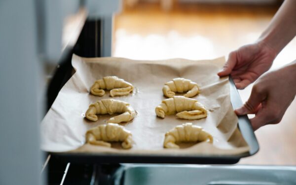 Hands placing homemade croissants on a tray into an oven, ready to bake.