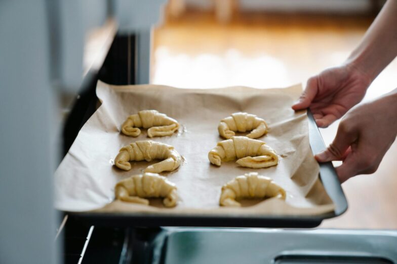 Hands placing homemade croissants on a tray into an oven, ready to bake.