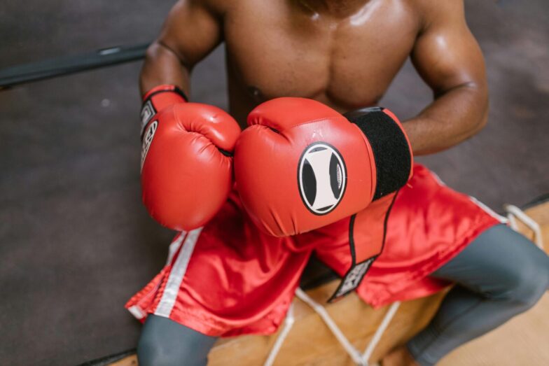Shirtless boxer in red gloves and shorts preparing for a match in the gym.