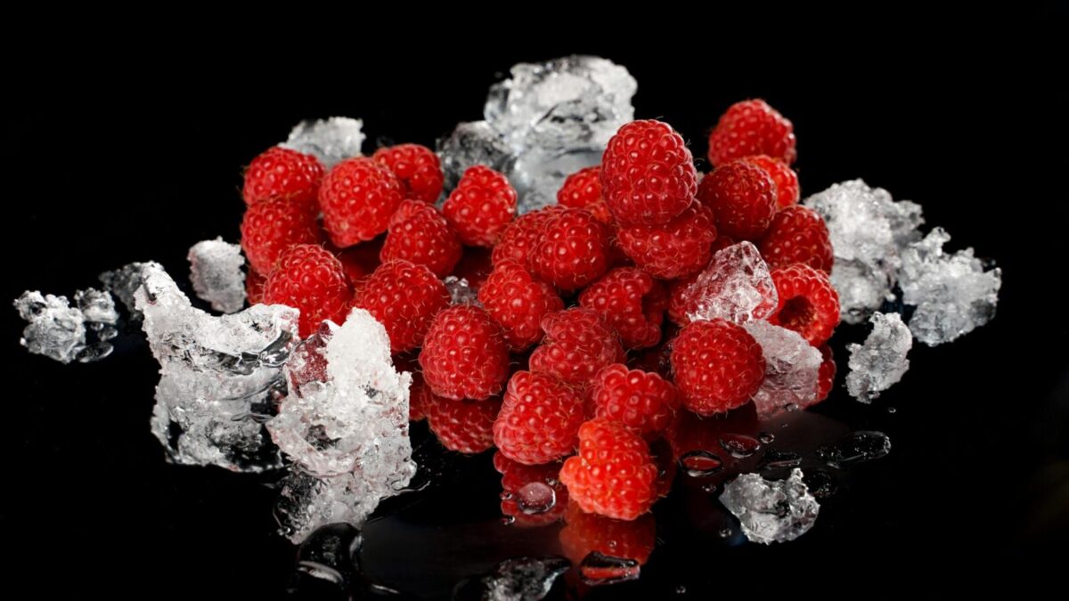 Close-up of vibrant red raspberries and melting ice on a reflective black surface.