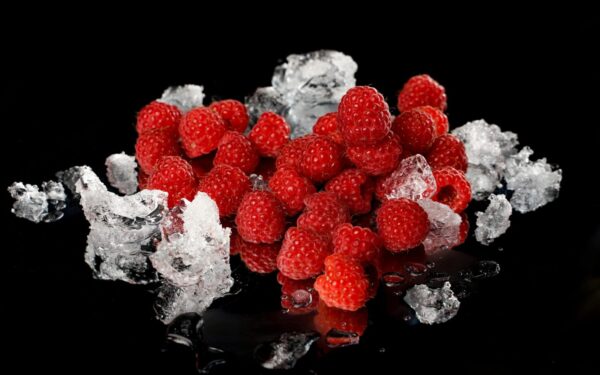 Close-up of vibrant red raspberries and melting ice on a reflective black surface.