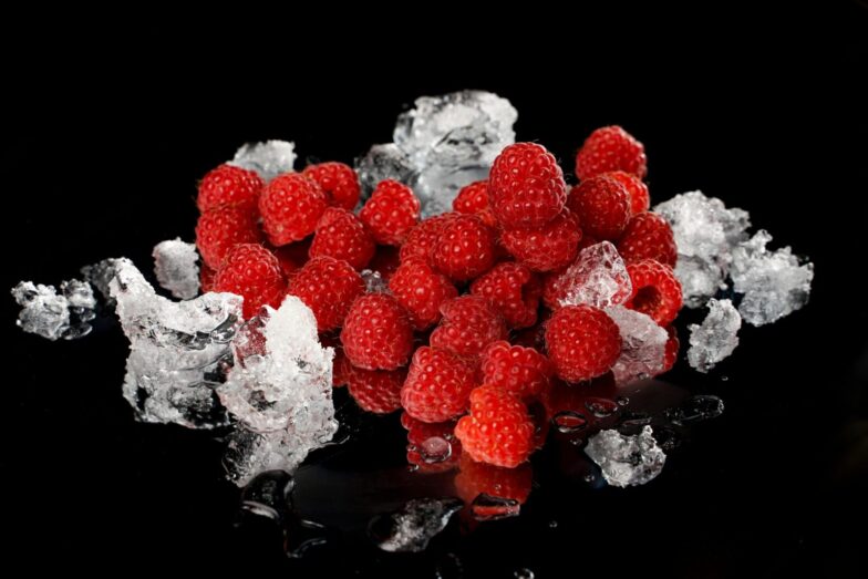 Close-up of vibrant red raspberries and melting ice on a reflective black surface.