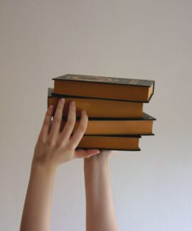 Hands reaching upwards holding a stack of hardbound books against a plain background.