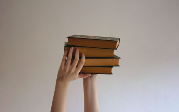 Hands reaching upwards holding a stack of hardbound books against a plain background.