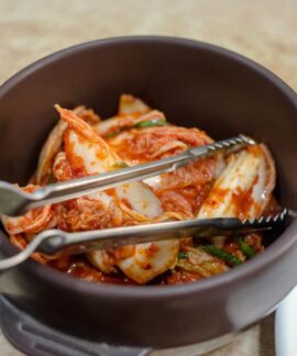 Close-up of traditional Korean kimchi in a dark ceramic bowl with tongs.